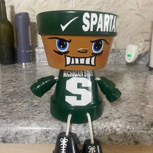 May include: A handcrafted terracotta pot transformed into a Spartan figure. The pot features a green and white colour scheme, with the word "SPARTANS" and a checkmark on the rim. The figure has blue eyes, a smiling face, and a green jersey with a white "S".