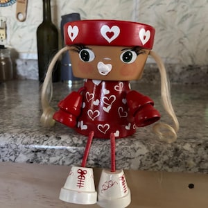 Puede incluir: Muñeca de terracota hecha a mano con un vestido y sombrero rojos, ambos adornados con corazones blancos. La muñeca tiene cabello rubio, ojos grandes y está diseñada para sentarse sobre una superficie. Las piernas de la muñeca son palos rojos y los pies son pequeñas macetas blancas.