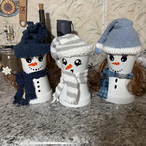 Pode incluir: Três bonecos de neve feitos de copos de plástico branco com rostos pintados, narizes de cenoura laranja e olhos de botão pretos. Cada um usa um gorro e cachecol de malha felpuda em azul marinho, listras cinzas e brancas ou azul claro.