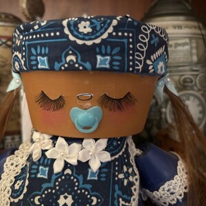 Puede incluir: Muñeca artesanal con cabeza de maceta de terracota, pintada con ojos cerrados, largas pestañas y mejillas rosadas. Lleva un pañuelo azul, un babero con detalles florales blancos y un chupete azul. Cabello castaño peinado en coletas.
