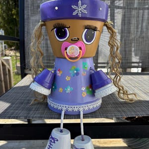 Op de afbeelding: Handgemaakte bloempotpop met een bruin gezicht, grote ogen, een fopspeen en krullend blond haar. Ze draagt een paarse jurk met bloemen en heeft bijpassende paarse armen en benen. De voeten van de pop zijn witte potten met paarse veters.