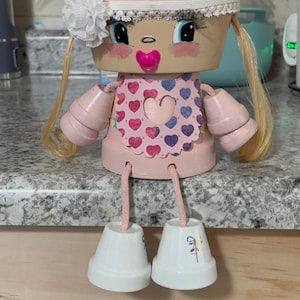 Puede incluir: Muñeca de maceta pintada a mano, en colores rosa y blanco. La muñeca tiene un babero rosa con diseños de corazones, pelo rubio y zapatos blancos. Cuenta con un chupete rosa y una flor blanca en la cabeza. Un artículo decorativo encantador.