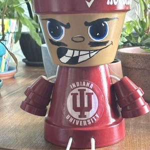 May include: A decorative terracotta pot figure painted in crimson and tan. The top section features a hand-painted face with blue eyes and a wide grin. The base displays the Indiana University logo. Two smaller pots serve as arms, attached with white cord.