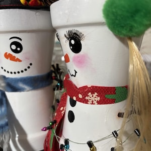Può includere: Due vasi bianchi a tema pupazzo di neve. Uno ha una sciarpa blu e un cappello a cilindro nero. L'altro ha una sciarpa rossa con un motivo a fiocco di neve, ciglia e un pompon verde. Entrambi hanno facce dipinte e luci decorative.