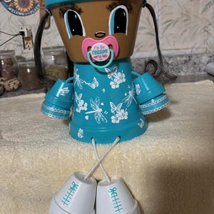 Puede incluir: Muñeca decorativa de maceta de terracota pintada a mano. Presenta una cara marrón, ojos grandes y un chupete. Viste un vestido turquesa adornado con mariposas y libélulas blancas. Las piernas son dos pequeñas macetas blancas con marcas turquesas.