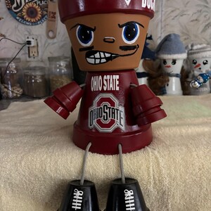 Puede incluir: Muñeca de maceta de terracota hecha a mano pintada con una cara marrón, ojos azules y un uniforme de fútbol rojo de Ohio State. La muñeca tiene un casco rojo con la palabra "BUCK" y una marca de verificación. Las piernas de la muñeca son macetas negras con marcas blancas de campo de fútbol.