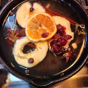 May include: A cast iron skillet filled with a dark liquid, featuring apple slices, an orange slice, cranberries, and star anise. Sprigs of rosemary and whole peppercorns add to the arrangement. A cinnamon stick is also visible.