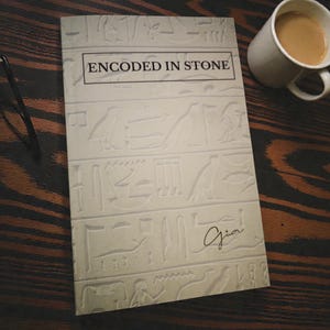May include: A book titled "ENCODED IN STONE" with hieroglyphic-style illustrations and the signature "Gia" on the cover. A mug of coffee sits beside the book on a dark wooden surface. The book's cover is off-white.