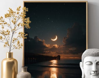 May include: A framed print depicts a nighttime coastal scene with a crescent moon, stars, and a pier extending into the ocean. The artwork is in a gold frame, with a gold vase and a stone Buddha head in the foreground.
