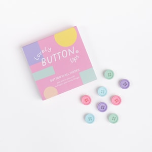 May include: A box of pastel-colored button wall hooks with the text "Lovely BUTTON Ups". The hooks are in shades of pink, blue, green, and purple. The box is pink with pastel accents.