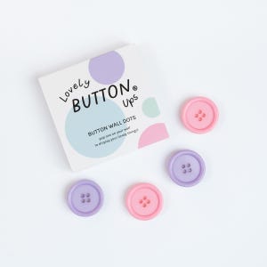 May include: A set of pastel-colored button wall dots. The package reads "Lovely BUTTON Ups" and "BUTTON WALL DOTS". Three purple and two pink buttons are scattered around the package. The buttons have four holes each.