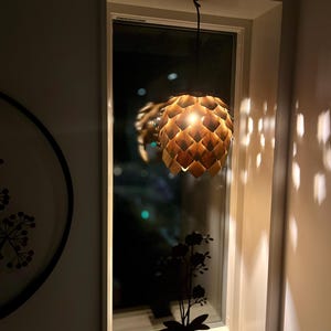 May include: A wooden pendant light with a geometric design hangs in front of a window. The warm light casts shadows on the wall. A decorative silhouette of a potted plant sits on the windowsill.