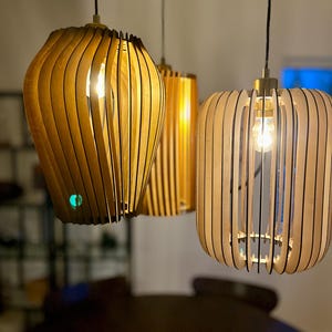 May include: Three wooden pendant lights with a warm, natural finish hang from the ceiling. Each light fixture features a unique design with vertical wooden slats, revealing the light bulbs inside. The lights are suspended by black cords.
