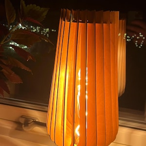 May include: A wooden table lamp with an orange, slatted shade. The lamp has a bulb that is lit, casting a warm glow. The lamp is on a windowsill, with a dark background.