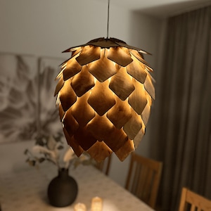 May include: A distinctive pendant light with a layered, geometric shade crafted from warm-toned wood, reminiscent of a pinecone. The light hangs from a single cord, emitting a soft glow. The fixture is suspended above a dining table, creating a warm ambiance.