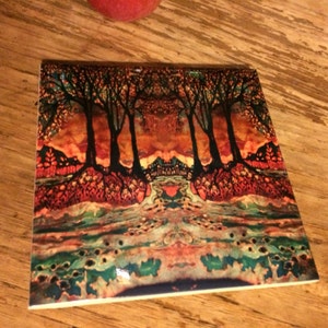 May include: A colorful ceramic tile with a painting of a forest scene. The trees are dark brown and the background is a mix of orange, red, and green.