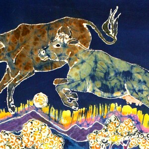 Puede incluir: Una pintura batik que representa dos vacas marrones y azules sobre un fondo azul con acentos amarillos, naranjas, morados y blancos. Las vacas est&aacute;n en una pose de salto, con una vaca en primer plano y la otra en el fondo.
