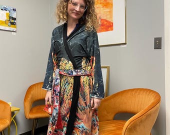 May include: A woman wearing a long, colorful robe with a black sash. The robe has a floral and abstract print. She is wearing yellow leggings and pink shoes. She is standing in front of a wall with two orange chairs and a painting.