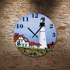 May include: Round wall clock featuring a scenic image of a white lighthouse with a black top, red-roofed buildings, and a blue sky. The clock face has black numerals and hands, and is set against a brick wall background.