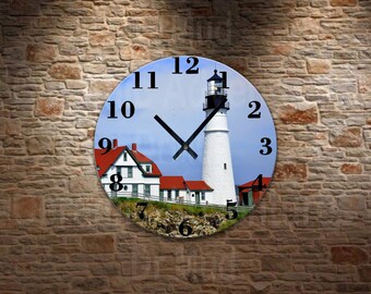May include: Round wall clock featuring a scenic image of a white lighthouse with a black top, red-roofed buildings, and a blue sky. The clock face has black numerals and hands, and is set against a brick wall background.