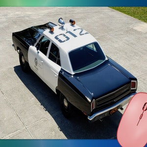 May include: A vintage black and white police car with the number "012" on the roof. The car has a white roof and doors, with the words "POLICE" and a badge visible. The car is parked on a concrete surface.
