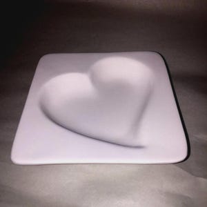 May include: A square, white ceramic plate with a heart-shaped indentation in the center. The plate has rounded corners and a smooth surface. The heart is subtly raised, creating a three-dimensional effect.