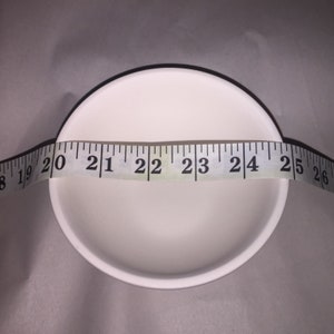 May include: A white ceramic bowl with a black rim is measured with a measuring tape. The bowl's diameter is approximately 22 inches. The bowl is empty and sits on a neutral-toned surface.