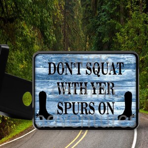 May include: A rectangular hitch cover with a blue wood grain background and the text "DON'T SQUAT WITH YER SPURS ON" in black. Black cowboy boot silhouettes are on each side of the text. The hitch cover is mounted on a black metal frame.