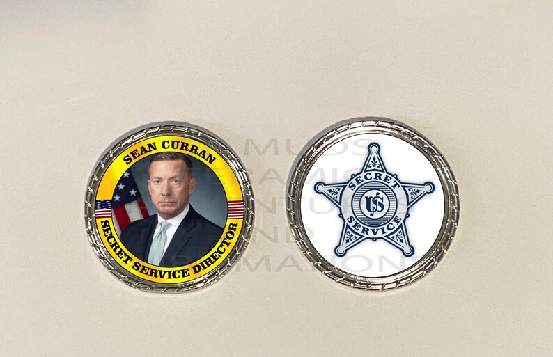 May include: Two silver-colored coins. One coin features a portrait of Sean Curran, Secret Service Director, with the American flag in the background. The other coin has a blue and white Secret Service star emblem.