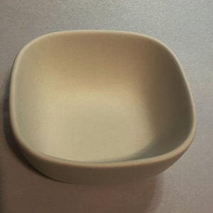 May include: A small, square, off-white ceramic bowl with rounded corners. The bowl has a smooth, matte finish and a simple, minimalist design. It is empty and ready to be filled.