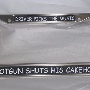 May include: A chrome license plate frame with the text "DRIVER PICKS THE MUSIC" at the top and "SHOTGUN SHUTS HIS CAKEHOLE" at the bottom. The text is in white on a black background.