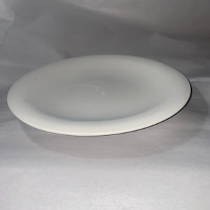 May include: A white, oval-shaped ceramic plate. The plate has a slightly raised rim and a smooth, glossy surface. The plate is sitting on a white surface.