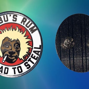 May include: A round pin with the text "JOBU'S RUM VERY BAD TO STEAL" encircling a cartoon face with a cigarette. The face has yellow hair and is set against a red background. The pin's back has two pin clasps.