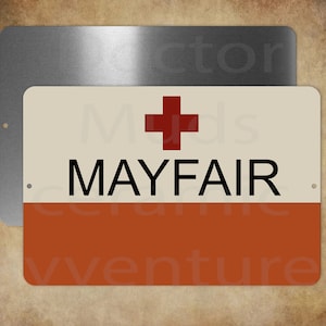 May include: A rectangular sign with the word "MAYFAIR" in black, a red cross, and a cream and orange color scheme. A second, plain, silver sign is behind the main sign. The sign has two small holes for mounting.