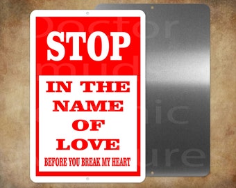 May include: A rectangular metal sign with a red border and a white center. Bold red text reads "STOP" at the top, followed by "IN THE NAME OF LOVE" and "BEFORE YOU BREAK MY HEART." A second, unadorned metal sign is visible.