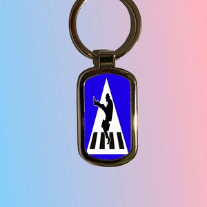 May include: Silver-colored metal keychain with a rectangular pendant featuring a blue background. The pendant displays a black silhouette of a person crossing a crosswalk, with a white triangle and black and white stripes.