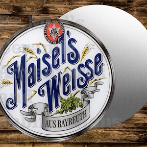 May include: Round beer coaster with the words "Maisel's Weisse" in blue script. The coaster has a white background with wheat stalks and a banner that reads "Aus Bayreuth". A second, plain silver coaster is next to it.