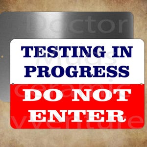 May include: A rectangular metal sign with the words "TESTING IN PROGRESS" in navy blue and "DO NOT ENTER" in white on a red background. The sign is mounted on a larger, brushed metal plate.
