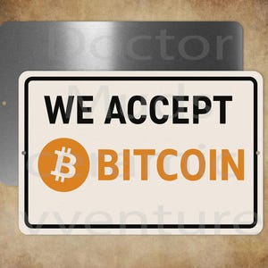 May include: A rectangular sign with the text "WE ACCEPT BITCOIN" in black and orange. The Bitcoin symbol is in an orange circle. The sign is white with a black border and is mounted on a brushed metal background.