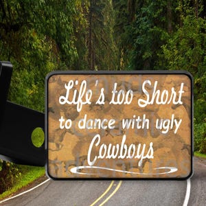 May include: Rectangular sign with the text "Life's too Short to dance with ugly Cowboys" in white script. The sign has a brown and tan background and a black border. The background shows a road and trees.