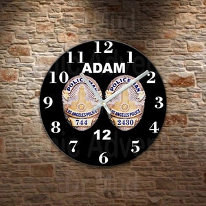 May include: A black wall clock featuring the name "ADAM" above two Los Angeles Police Department badges. The clock face displays the time with white numerals and hands. The badges have the numbers 744 and 2430.