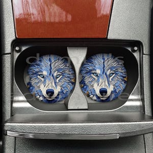 May include: Two round car coasters featuring a detailed wolf face design. The wolf's fur is depicted in shades of blue and white, with piercing blue eyes. The coasters are set in a car's cup holder.