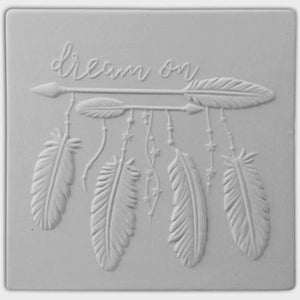 May include: A square, light gray decorative tile with an embossed dreamcatcher design. The design includes an arrow, feathers, and the words "dream on" in cursive script. The feathers hang from the arrow with small star-shaped beads.