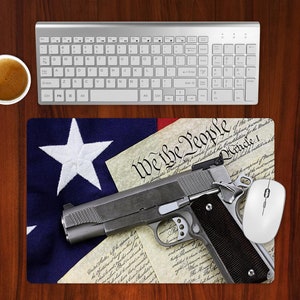 May include: A computer desk setup featuring a silver keyboard, a white mouse, and a mousepad with an American flag design and the text "We the People." A handgun rests on the mousepad. A cup of coffee sits to the left.