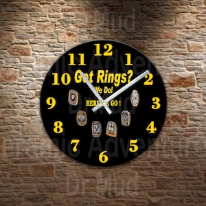 May include: Black round wall clock with yellow numerals and hands. The clock face features the text "Got Rings? We Do! HERE WE GO!" and images of championship rings. The clock is mounted on a brick wall.