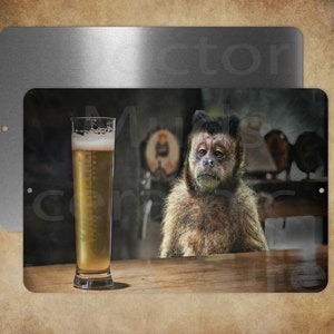 May include: Metal sign featuring a monkey sitting at a bar with a tall glass of beer. The monkey has a contemplative expression. The background includes a tap and bar fixtures. The sign has a distressed, vintage look.