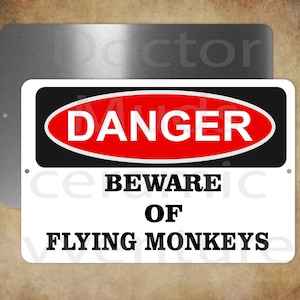 May include: A rectangular metal sign with the words "DANGER" in a red and black oval, and "BEWARE OF FLYING MONKEYS" in bold black text. A second, plain metal sign is behind it.