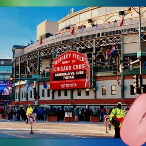 May include: Exterior view of Wrigley Field, home of the Chicago Cubs, with a red sign displaying the game details: Cardinals vs. Cubs at 7:05 P.M. The stadium has a classic design with a brick facade and a green outfield. People are gathered outside.