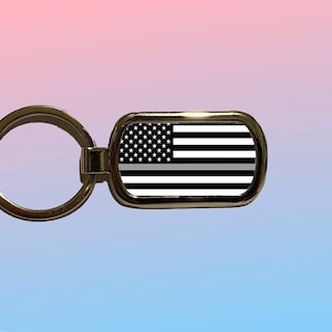 May include: A rectangular keychain with a gold-colored frame. The keychain features a design of the American flag in black and white, with a gray stripe. The keychain is attached to a gold-colored key ring.