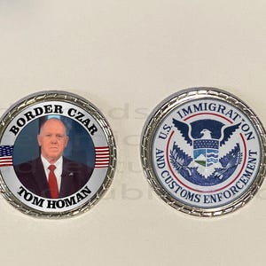 May include: Two silver-colored coins. One coin features a portrait of Tom Homan, with the text "BORDER CZAR" and "TOM HOMAN" around the edge, along with an American flag. The other coin displays the U.S. Immigration and Customs Enforcement seal.